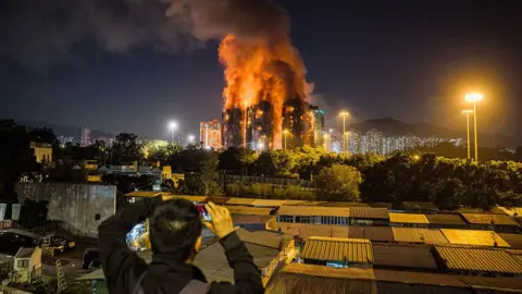 AFP via Getty Images A man takes a photo of the huge fire, which is in the distance but dominating the skyline