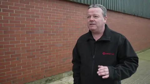 Nick Harman, 56, who wears a black zip-up fleece and has grey hair, speaks to the camera. 