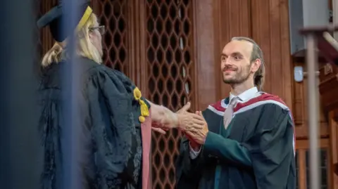 University of Bristol Paul Edwards, who has blonde hair and a beard, wearing gown and standing in hall where he graduated.