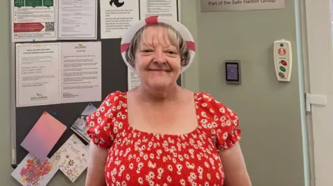 A woman with a white and red England hat and wearing a red dress with a white-flower pattern, smiles as she stands in front of a notice board.