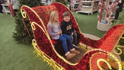 A young girl with blonde hair is sitting next to a young boy with brown hair, he has a breathing tube going into his nose. They are sitting in a large sleigh which is red and covered in gold lights.