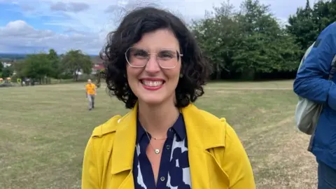 A woman is smiling. She is wearing glasses and yellow jacket.