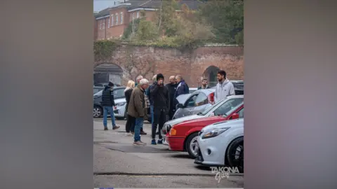 Lewis Warren Group of people gathered in a car park beside several parked vehicles, including a red car and a silver car in the foreground. Brick walls with greenery line the background, and one person is holding a cup while others stand and talk. A “TAKONA Crew” watermark appears in the bottom right corner.
