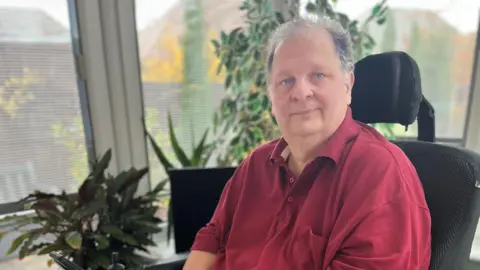 Marcus Watkins A man wearing a red polo shirt sat in a black powered wheelchair inside a conservatory, with plants inside.