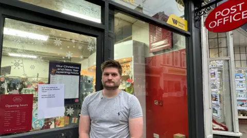 A blonde man wearing a light grey t-shirt standing outside the glass windows of a post office. On the door next to him is a sign explaining the post office is closed because of a break-in.