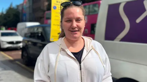 A woman with dark hair and sunglasses on her head, she has a white coat and dark top