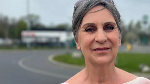 A woman wearing a pink-coloured top looks towards the camera while standing in front of a roadside services 