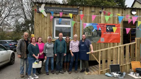 BBC Volunteers and members of the team stood in a line infront of the shop which is clad in wood. 