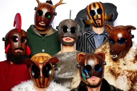 Lionel Pesqué The band FÜLÜ are wearing strange wooden masks, including ones that looks like an owl, a bear and a wolf with big teeth. The band members are standing together and pictured against a white background.