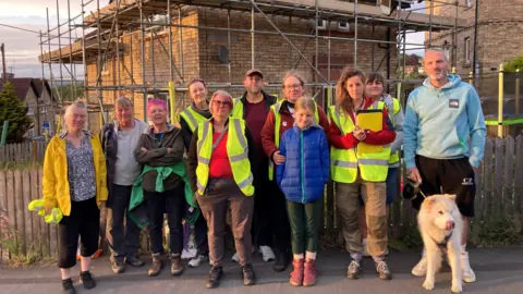 Save Newcastle Wildlife Residents and members of Save Newcastle Wildlife in hi-vis vests standing in front of a brown brick house covered in scaffolding on Monday evening. One of the residents is holding a white fluffy dog on a lead.