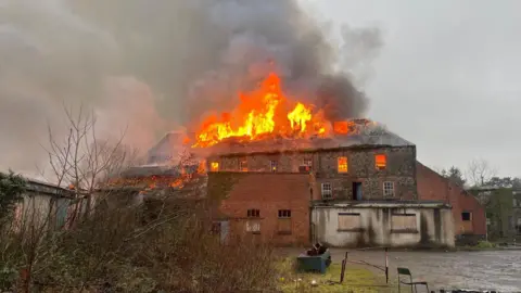 A red and grey derelict building, with large flames coming from the roof. Plumes of dark grey smoke are rising from the building. 