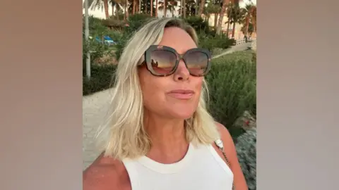 Selfie of Becky Curtis, wearing a white vest top and large, brown sunglasses, while on holiday with palm trees in the background