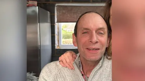 Andy Reed. He has dark thinning hair and stubble. He is looking at the camerta and smiling. He is pictured wearing a three-quarter zip light brown jumper. The hand of a woman who cannot be seen is on his right shoulder.