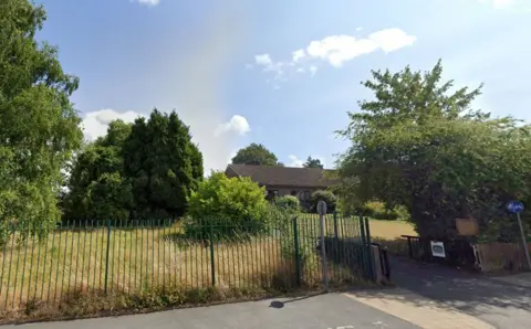 Google A building beyond a grassy field and lawn seen behind railings from the street. It is a sunny day. The building appears to be single-storey.