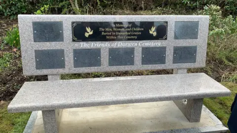 A stone bench is set on a concrete slab. It has a number of plaques on it. The one at the top reads: 'In memory of the men, women and children buried in unmarked graves within this cemetery'