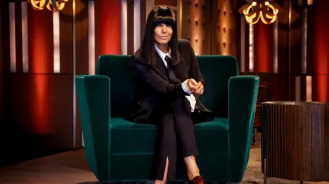 Claudia Winkleman sits on a green velvet sofa wearing a burgundy blazer and trousers