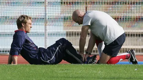 Getty Images Jonny Wilkinson lying on the ground receiving treatment on an injured ankle from physio Phil Pask. Pask is putting pressure on his foot and is wearing a white T-shirt and blue shorts. 