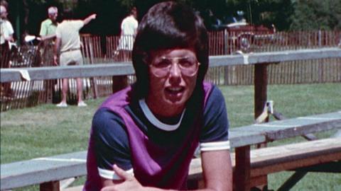 Billie Jean King in a purple and blue T-shirt, sat on bleachers being interviewed.  In the background, a few people conversing at a fence.