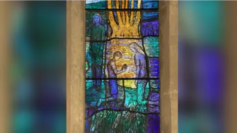 Image of a stained glass window depicting a boy who is kneeling down looking at a shell.  A young girl is standing over him while a second girl looks on.