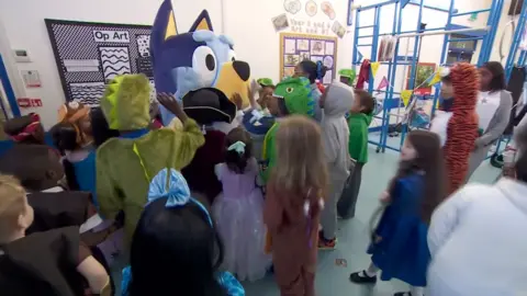 A group of children in a school hall gathered around Bluey in character