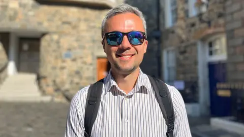 A man with light grey hair smiling at the camera. He is wearing sunglasses and has a pale pink shirt with blue pinstripes. You can see the straps of a grey rucksack over his solders.
