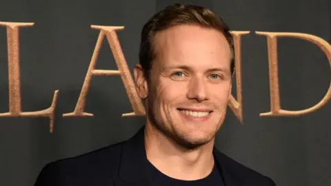 Sam Heughan looking straight at the camera while standing in front of an Outlander backdrop.