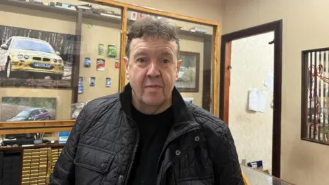 A man with a black coat, black t-shirt, and short brown hair stands in a shop. There is a picture of a yellow car on the wall behind him.
