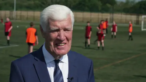 An older man, with white hair and wearing a navy blue suit, speaks to the camera at the side of a football pitch