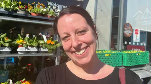 A smiling woman stood in front of flowers and plants.