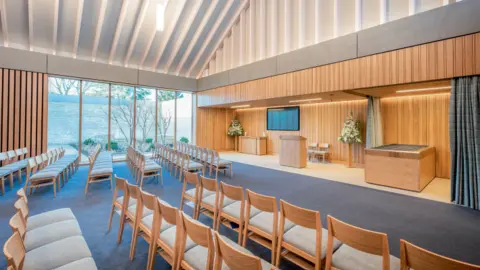 The interior of a large room at Guildford Crematorium. Rows of wooden chairs can be seen pointing towards an altar-type area. There are long panelled windows, a blue carpet and a television screen on the wall.