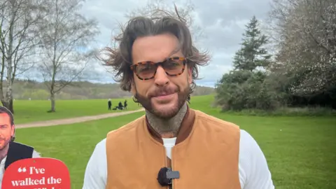 Alicia Owen/BBC Pete Wicks has a big floppy brown fringe which is windswept, thick framed glasses and is wearing a brown vest jacket. Behind him is the grass lawns of the country park and trees.