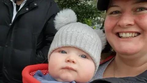 Family Emma is smiling and holding Theo in front of her - he's wearing a grey hat and red jumper