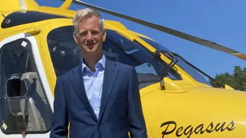 Dorset and Somerset Air Ambulance Charles Hackett is wearing a blue suit and standing in front of a yellow helicopter.