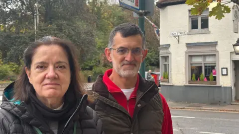 Carolyn Thornborrow, wearing a hooded winter coat and Duncan Jack, wearing a padded gilet and a red-zipped jumper underneath, stand in Station Street, Quorn, which was submerged by flood waters in January. A sign for the corner of High Street can be seen on the side of a pub in the background. 