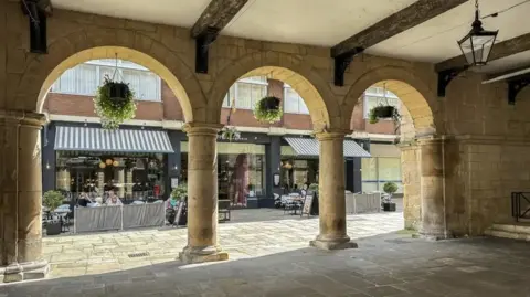 Getty Images View across the Square in Shrewsbury town centre from the Old Market Hall. There is a paved area and the building has arches and pillars. Across the square, people are sitting at tables and there are shop awnings.