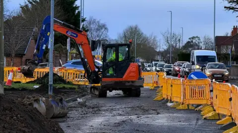 A road digger in operation on a sectioned off lane of a road with yellow barriers around it. Cars queue up on the lane next to it.