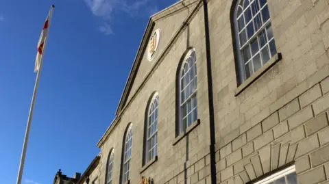 BBC A Guernsey flag and blue sky stands next to a building with grey bricks and blue windows, showing the States logo. 
