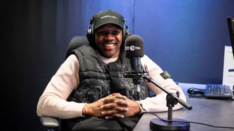 Rapper Giggs is sat smiling in a BBC 1Xtra radio studio. He is wearing a black cap with a black body warmer and white jumper with a microphone in front of him.