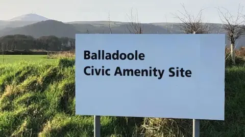 A white sign that reads Balladoole Civic Amentiy Site in front of a field with hills in the background.