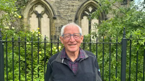 Jessica Bayley/BBC A man in his late 60's wearing a black quarter-zip fleece over a red and blue checked shirt and glasses smiles whilst stood in front of a steel fence in front two arches covered by greenery.