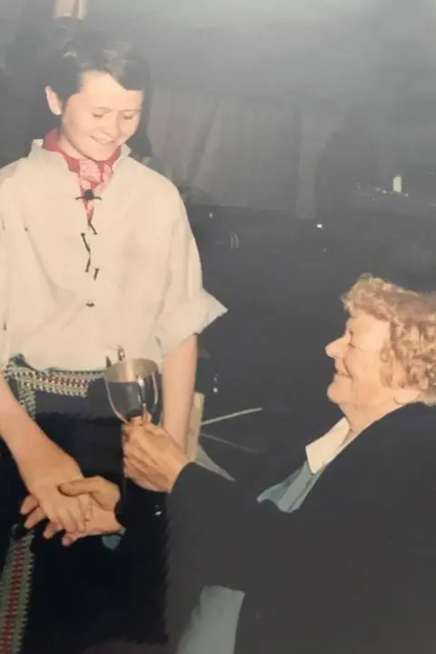 JUAN GARRETT A young Juan Garrett receiving a trophy. He is is wearing a white shirt and black trousers and is being handed the cup by an older lady with blonde hair, who is wearing a dark jacket.
