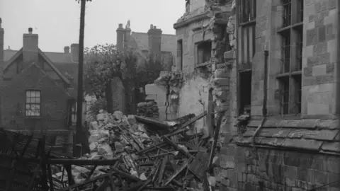 A black-and-white image showing rubble in the middle of the photo. One building is nearest to the camera on the right of the image. 