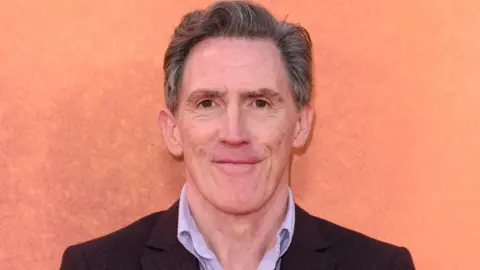 Headshot of Brydon against an orange background. He wears a brown blazer and lilac shirt underneath. 