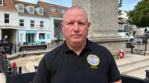 Darren is pictured in front of the cenotaph. He's wearing a black veterans polo shirt and has a serious look. He has a shaved head. 