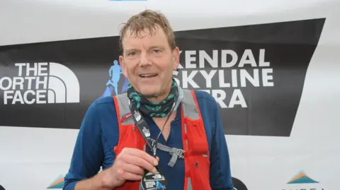 David Griffin David Griffin is standing in front of a poster for the Kendal Skyline Ultra and holding a medal. He is wearing a hi-vis vest and blue top and is a man in his 60s with brown hair 