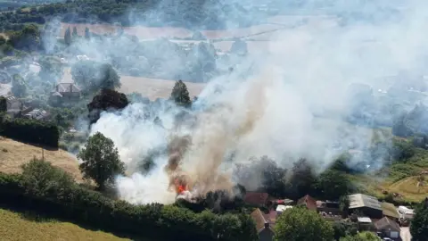Fire and smoke can be seen rising from a rural plot of land with houses surrounded by fields and trees