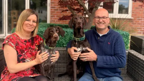 Duncan Horlor sat on a brown straw bench with his partner and two brown dogs. Duncan is bald, wears glasses a blue jumper and blue jeans while his partners has a blonde bob and smiles with her arms around one of the brown dogs. She is wearing a pink floral dress and black tights.