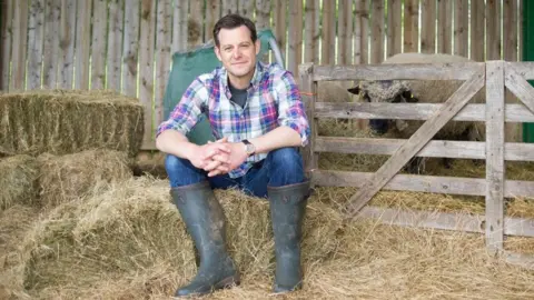 LAS Matt has short brown hair and is wearing a pink, blue and white checked shirt, blue jeans and grey wellies. He is sitting on hay bales in a barn in front of a wooden gate. Behind him is a sheep.