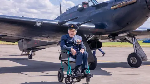 RAF John 'Paddy' Hemingway sits in a wheelchair on an airstrip close to an RAF jet. He is smiling at the camera as he wears a full uniform