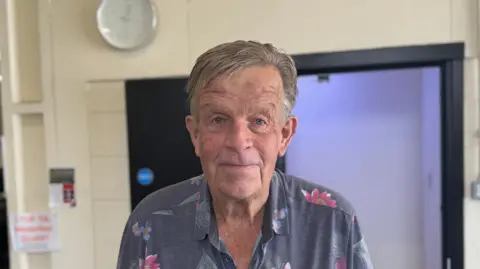 George King/BBC A head and shoulders image of Roger Mayhew. He is wearing a short sleeved shirt with flowers on it. He is looking directly into the camera.